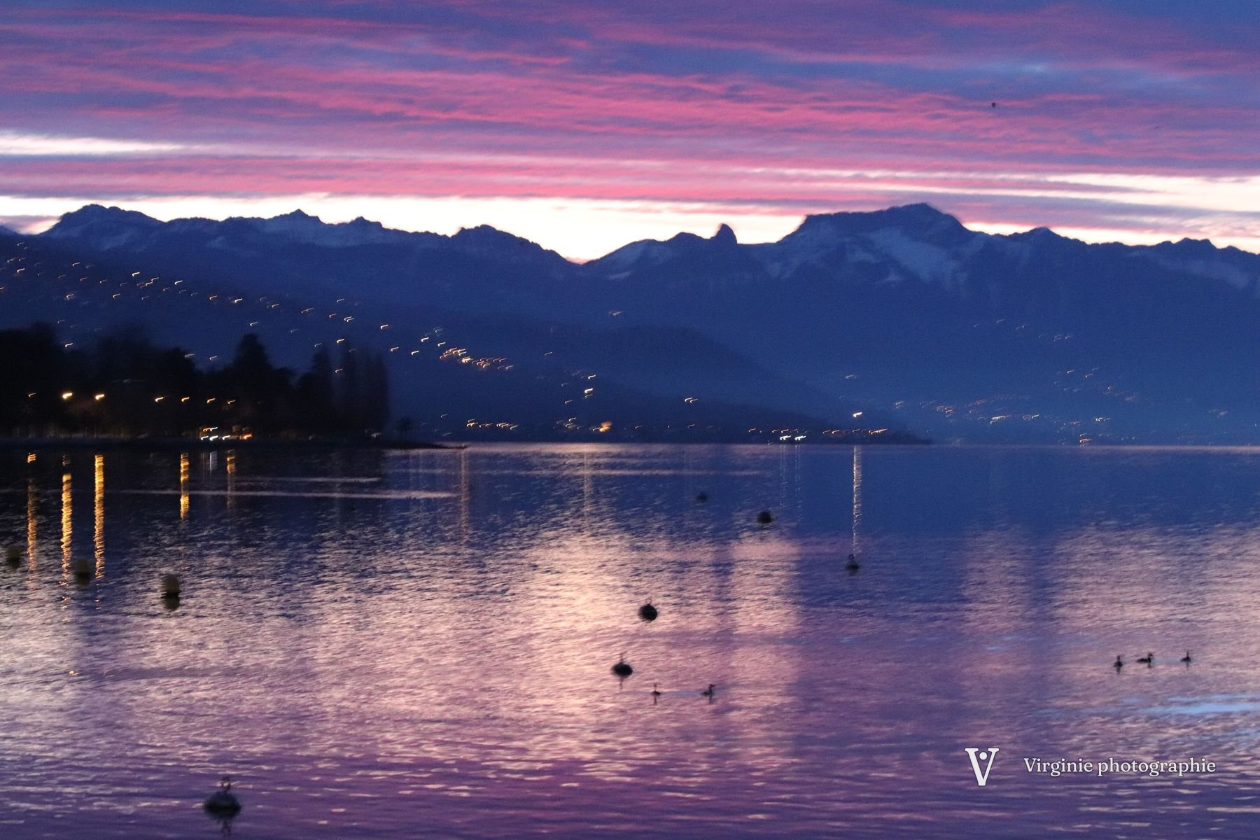 Le Léman au crépuscule, ciel rose et violet au-dessus des montagnes, les lumières de la rive se reflètent dans l’eau